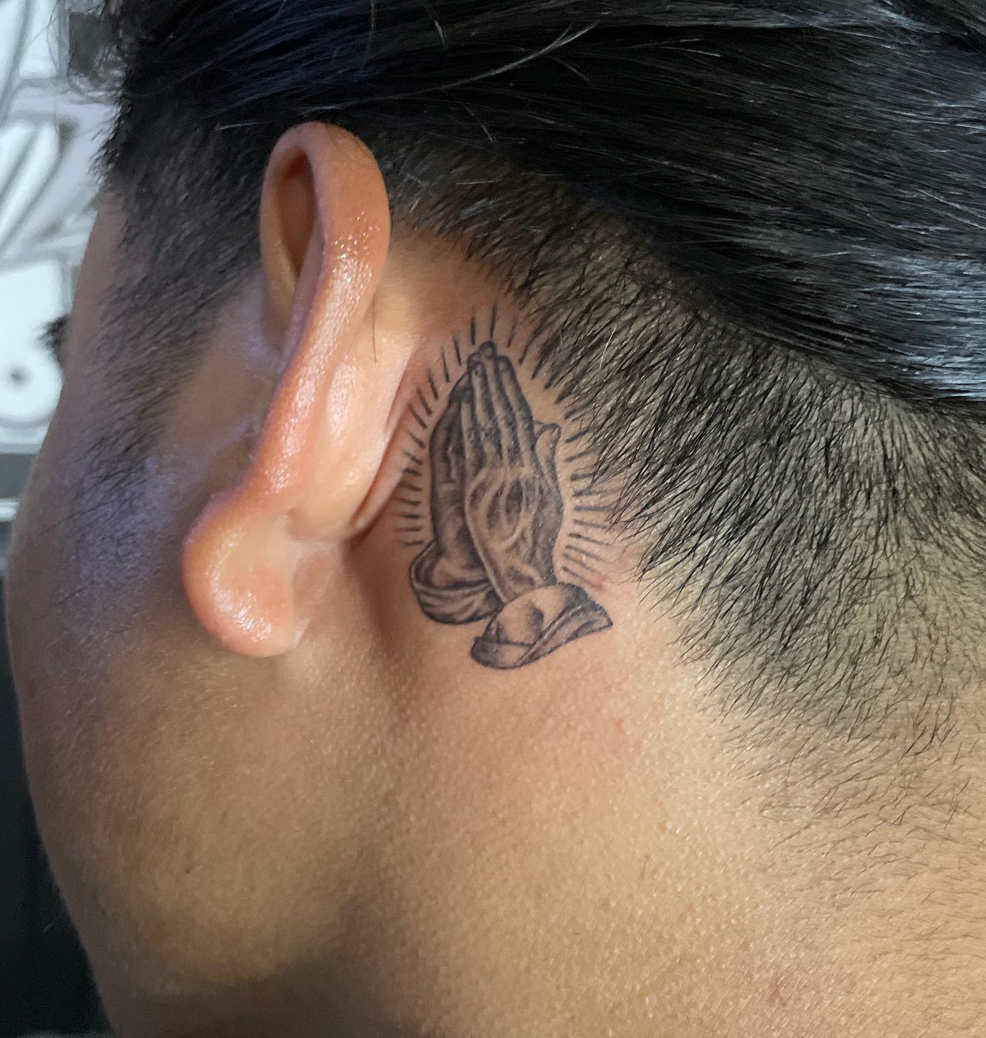 Tatuaje de manos rezando detrás de la oreja.